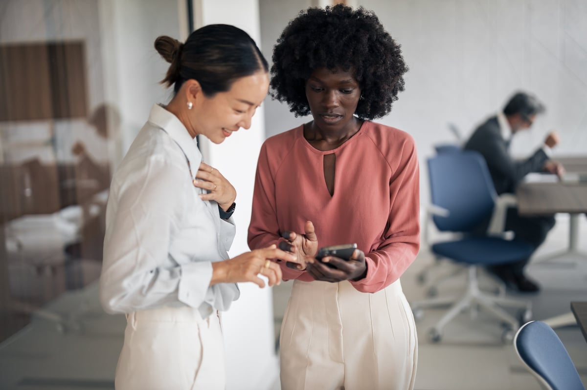 Diverse businesswomen are collaborating closely, looking at information on a smartphone screen together in a modern office environment, demonstrating teamwork and professional communication